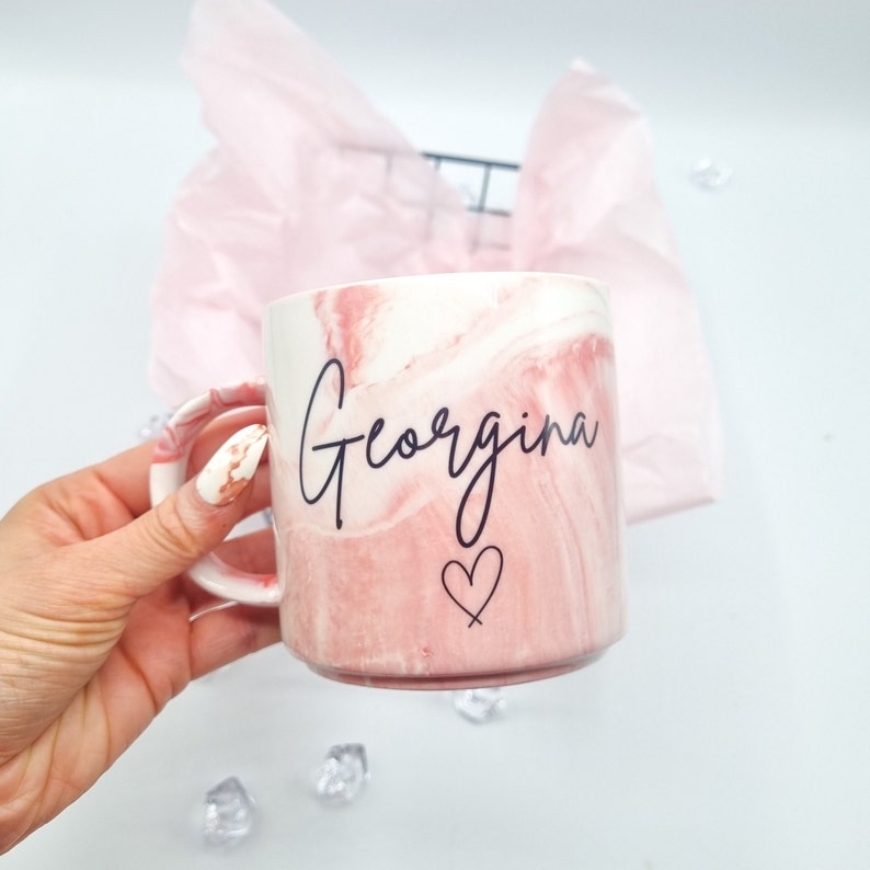 May include: A pink and white marble mug with the name "Georgina" written in black cursive script and a small heart symbol.