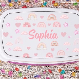 May include: A white lunchbox with a pink and rainbow design. The lunchbox features the name "Sophia" in pink script, along with pink hearts, stars, rainbows, and clouds. The lunchbox has a white lid and a purple base.