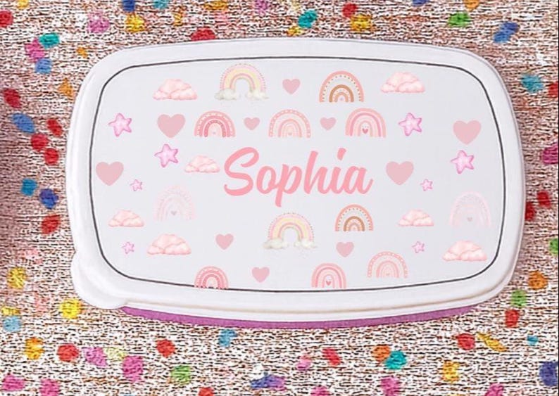 May include: A white lunchbox with a pink and rainbow design. The lunchbox features the name "Sophia" in pink script, along with pink hearts, stars, rainbows, and clouds. The lunchbox has a white lid and a purple base.