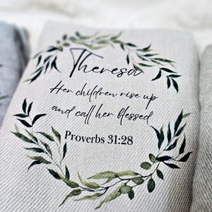 May include: A white canvas throw pillow with a green leafy wreath design. The pillow features the name "Theresa" and the quote "Her children rise up and call her blessed" with the scripture reference "Proverbs 31:28".