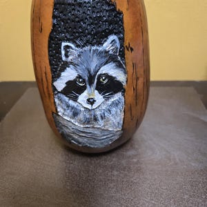 May include: A hand-carved gourd featuring a raccoon design. The raccoon is painted in shades of gray, black, and white, with detailed fur and expressive eyes. The gourd has a natural brown color with a carved-out area around the raccoon.