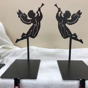 Puede incluir: Dos soportes de calcetines de metal negro en forma de ángel con una trompeta en sus manos. Los ángeles están en silueta sobre un fondo blanco.