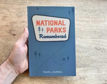 May include: A blue travel journal with a retro-style sign that says "National Parks Remembered" and a design of trees and a tent. The journal has the words "Travel Journal" printed on the bottom.