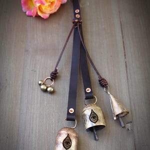 May include: A decorative leather strap with three metal bells and a cluster of small beads. The bells have an eye design. A pink and yellow rose rests on a wooden surface.
