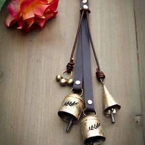 May include: A decorative leather strap with three gold-colored bells and a small cluster of brass bells. The bells have a leaf design and hang from a dark brown leather strap. A red and yellow rose is in the background.