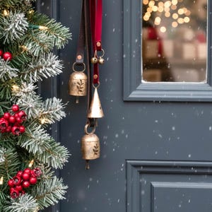以下が含まれることがあります： 茶色と赤色の革ストラップに吊るされた3つの真鍮ベルが特徴の、お祝いのクリスマスのドアの装飾。 ベルには花の模様があり、青いドアと雪の降る常緑樹の枝を背景に吊るされています。