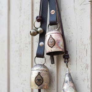 May include: A collection of rustic metal bells with leather straps. The bells vary in shape, including a cone and teardrop design, and feature an eye symbol. The bells are attached to dark brown leather straps, and are hanging against a white wooden background.