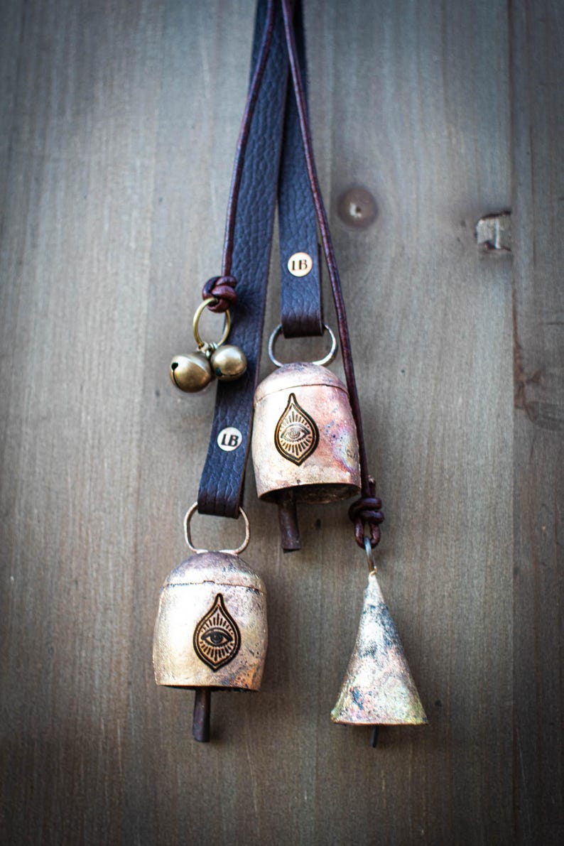 May include: Close-up of several metal bells hanging from dark brown leather straps. The bells vary in shape, including a teardrop and a cone. Each bell has a unique design, and the straps are attached with metal rings and knots. The background is a wooden surface.