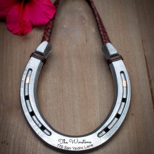 Op de afbeelding: Een zilveren hoefijzervormige wandhanger met een bruine leren riem. Het hoefijzer heeft de tekst "The Winstons 770 San Ysidro Lane" gegraveerd. Een rode hibiscusbloem is zichtbaar op de achtergrond.