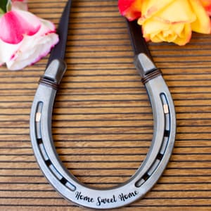 Op de afbeelding: Een zilveren hoefijzervormige wanddecoratie met de tekst "Home Sweet Home". Het hoefijzer heeft een bruine leren riem en is versierd met twee rozen, een roze en witte en de andere geel en oranje.