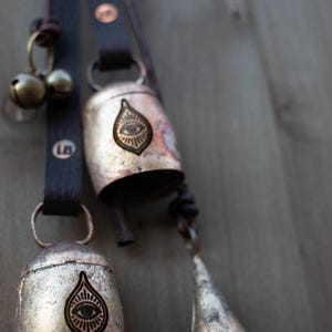 May include: Close-up of rustic metal bells with leather straps. The bells have an eye design and are attached to a dark brown leather strap. The bells are in varying sizes and shapes, with a small bell and a cone-shaped bell.