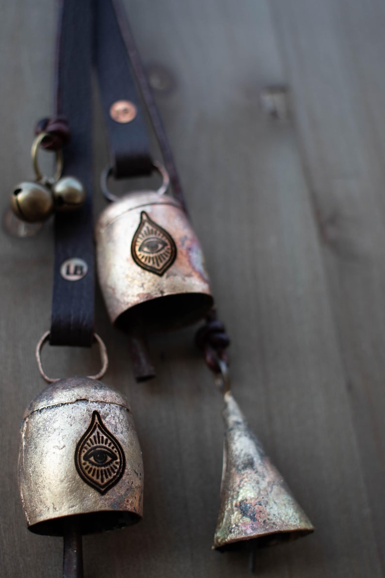 May include: Close-up of rustic metal bells with leather straps. The bells have an eye design and are attached to a dark brown leather strap. The bells are in varying sizes and shapes, with a small bell and a cone-shaped bell.