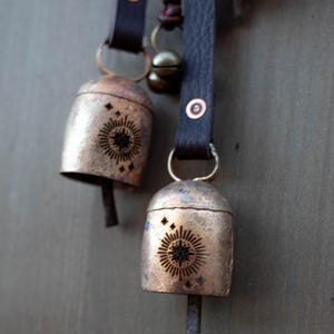 May include: Two rustic metal bells with a sunburst design and star accents hang from dark brown leather straps. The bells have a tarnished, aged appearance, and are attached with metal rings and rivets. A small brass bell hangs nearby.