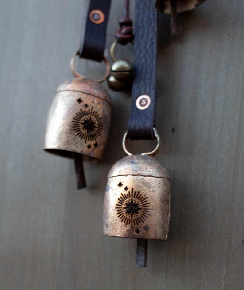May include: Two rustic metal bells with a sunburst design and star accents hang from dark brown leather straps. The bells have a tarnished, aged appearance, and are attached with metal rings and rivets. A small brass bell hangs nearby.