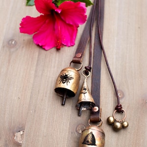 May include: A collection of brass bells with leather straps, featuring bee and beehive designs. A bright pink hibiscus flower and green leaves are in the background. The bells are of varying shapes and sizes.