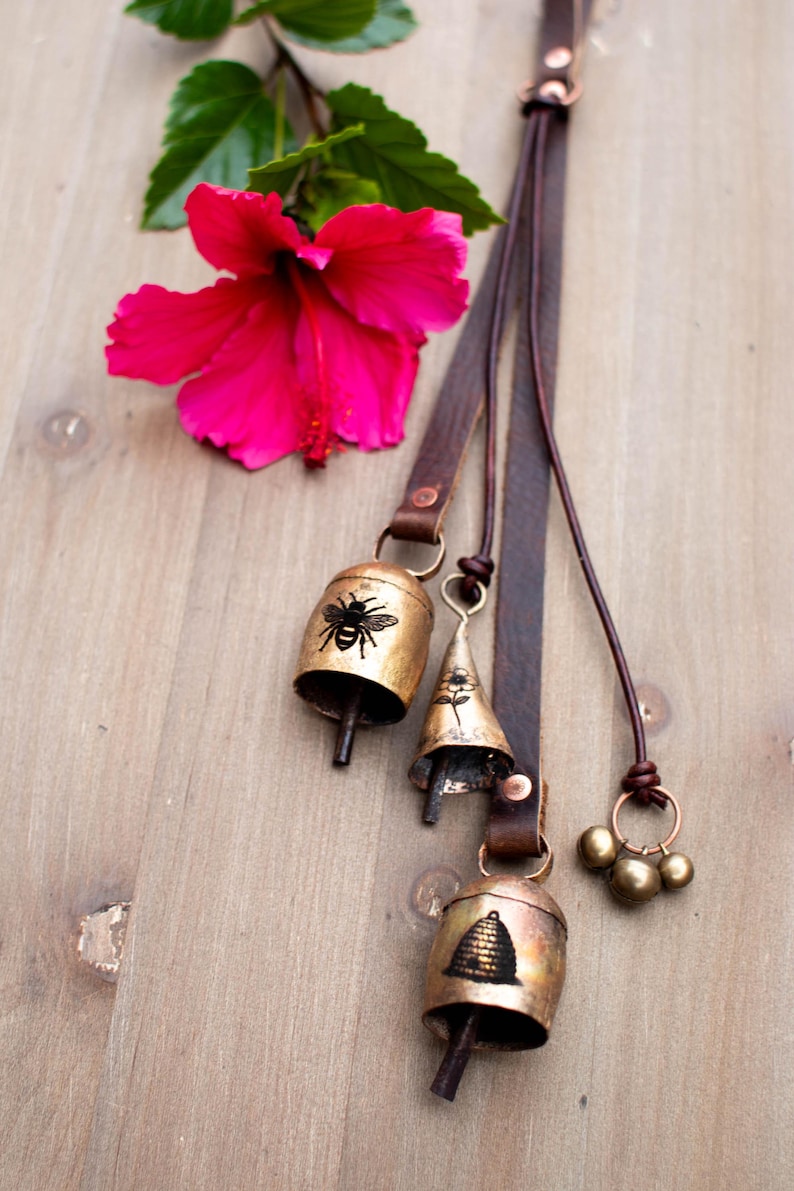 May include: A collection of brass bells with leather straps, featuring bee and beehive designs. A bright pink hibiscus flower and green leaves are in the background. The bells are of varying shapes and sizes.