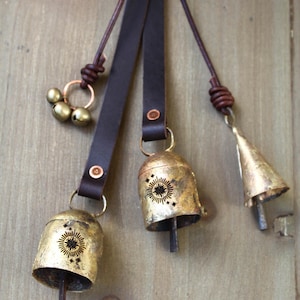 May include: A collection of rustic brass bells with leather straps. The bells vary in shape and size, some with a sunburst design. The leather straps are dark brown and attached with rivets. The bells are hanging against a wooden background.