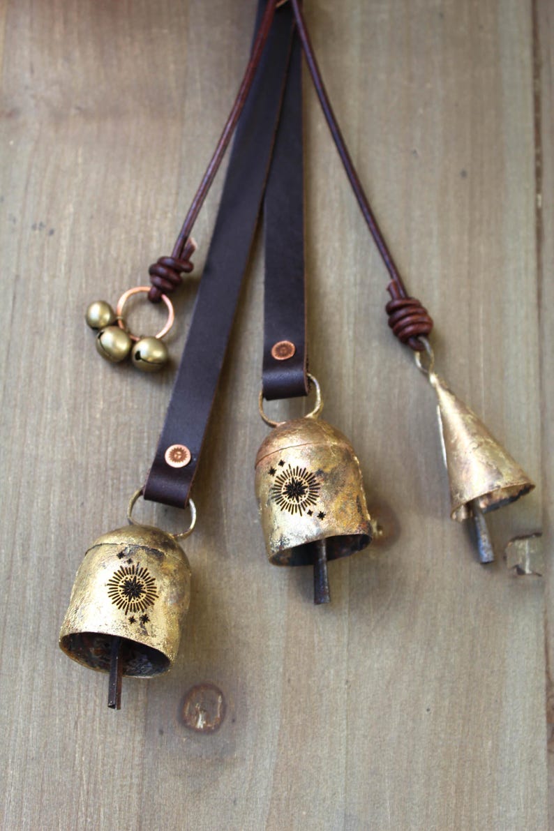 May include: A collection of rustic brass bells with leather straps. The bells vary in shape and size, some with a sunburst design. The leather straps are dark brown and attached with rivets. The bells are hanging against a wooden background.