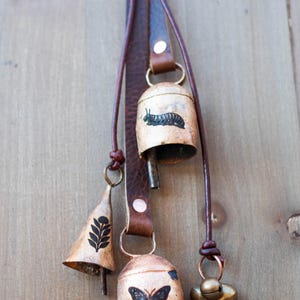 May include: A collection of rustic metal bells with leather straps. The bells feature various designs, including a caterpillar, a butterfly, and a leaf pattern. The bells are in varying shapes and sizes, with a warm, aged finish.