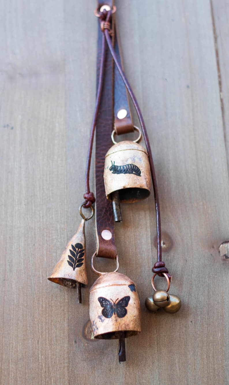 May include: A collection of rustic metal bells with leather straps. The bells feature various designs, including a caterpillar, a butterfly, and a leaf pattern. The bells are in varying shapes and sizes, with a warm, aged finish.