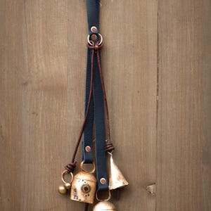 May include: A decorative hanging with three brass bells and a small brass ball, suspended from a black leather strap. The bells have a sunburst design and are attached with brown leather and copper rings. The background is a wooden surface.