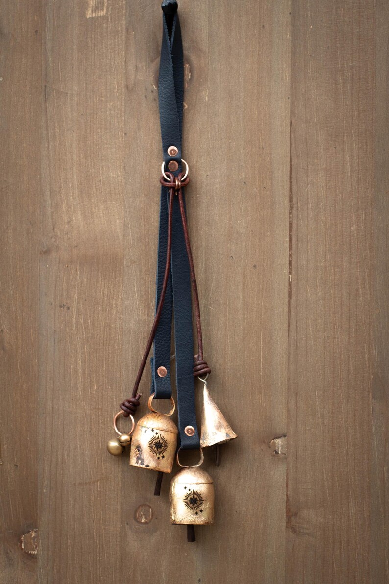 May include: A decorative hanging with three brass bells and a small brass ball, suspended from a black leather strap. The bells have a sunburst design and are attached with brown leather and copper rings. The background is a wooden surface.