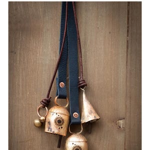 May include: Blue leather straps with brown round leather cords, holding several antique gold bells. The bells have a sunburst design and are hanging against a wooden background.