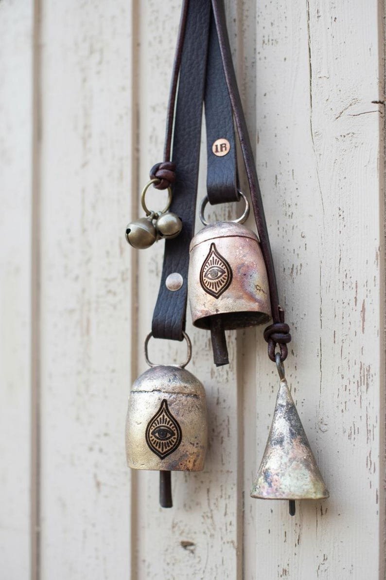 May include: A collection of rustic metal bells with leather straps. The bells vary in shape, including a cone and teardrop design, and feature an eye symbol. The bells are attached to dark brown leather straps, and are hanging against a white wooden background.