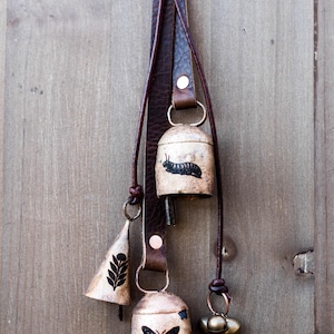 May include: A set of decorative bells hanging from a brown leather strap. The bells are various shapes and sizes, with a butterfly, caterpillar, and leaf design. The bells are made of metal and have a rustic, aged appearance.