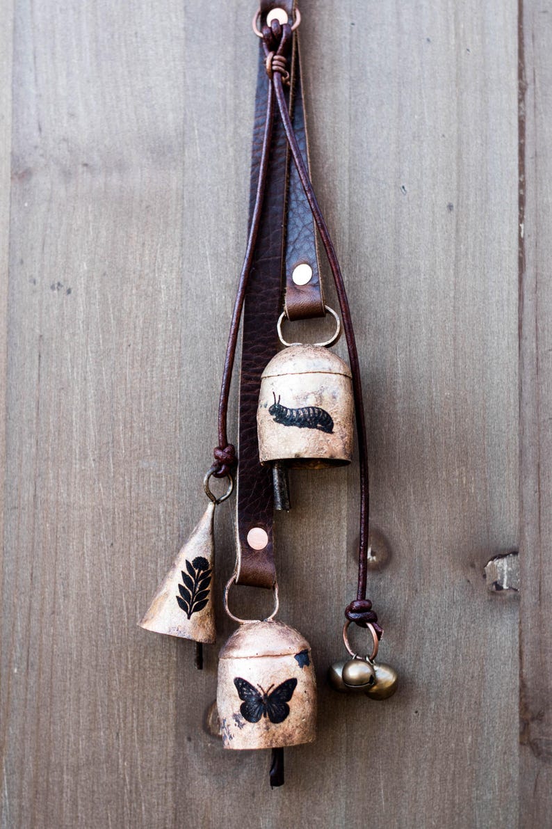 May include: A set of decorative bells hanging from a brown leather strap. The bells are various shapes and sizes, with a butterfly, caterpillar, and leaf design. The bells are made of metal and have a rustic, aged appearance.