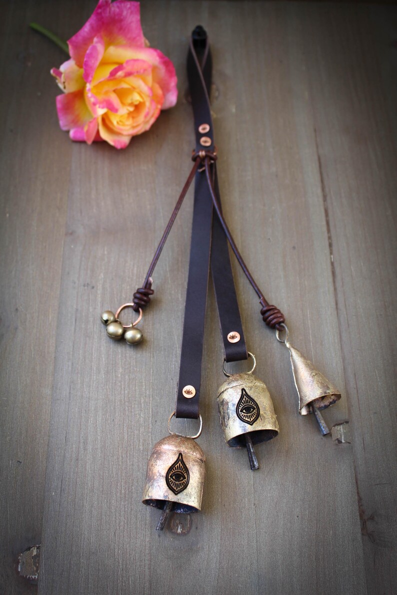 May include: A decorative leather strap with three metal bells and a cluster of small beads. The bells have an eye design. A pink and yellow rose rests on a wooden surface.