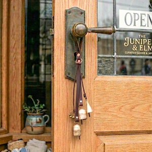 May include: A rustic wooden door with a bronze handle and a leather strap holding small bells. The door has a sign that says "OPEN" and the shop name "THE JUNIPER & ELM". A small potted plant sits in the window.