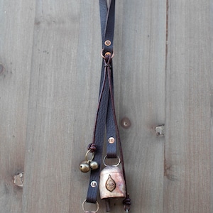 May include: A decorative hanging with three bells and two smaller brass bells. The bells are attached to a dark brown leather strap. The bells have a metallic finish with an eye design. The hanging is suspended from a brass hook.