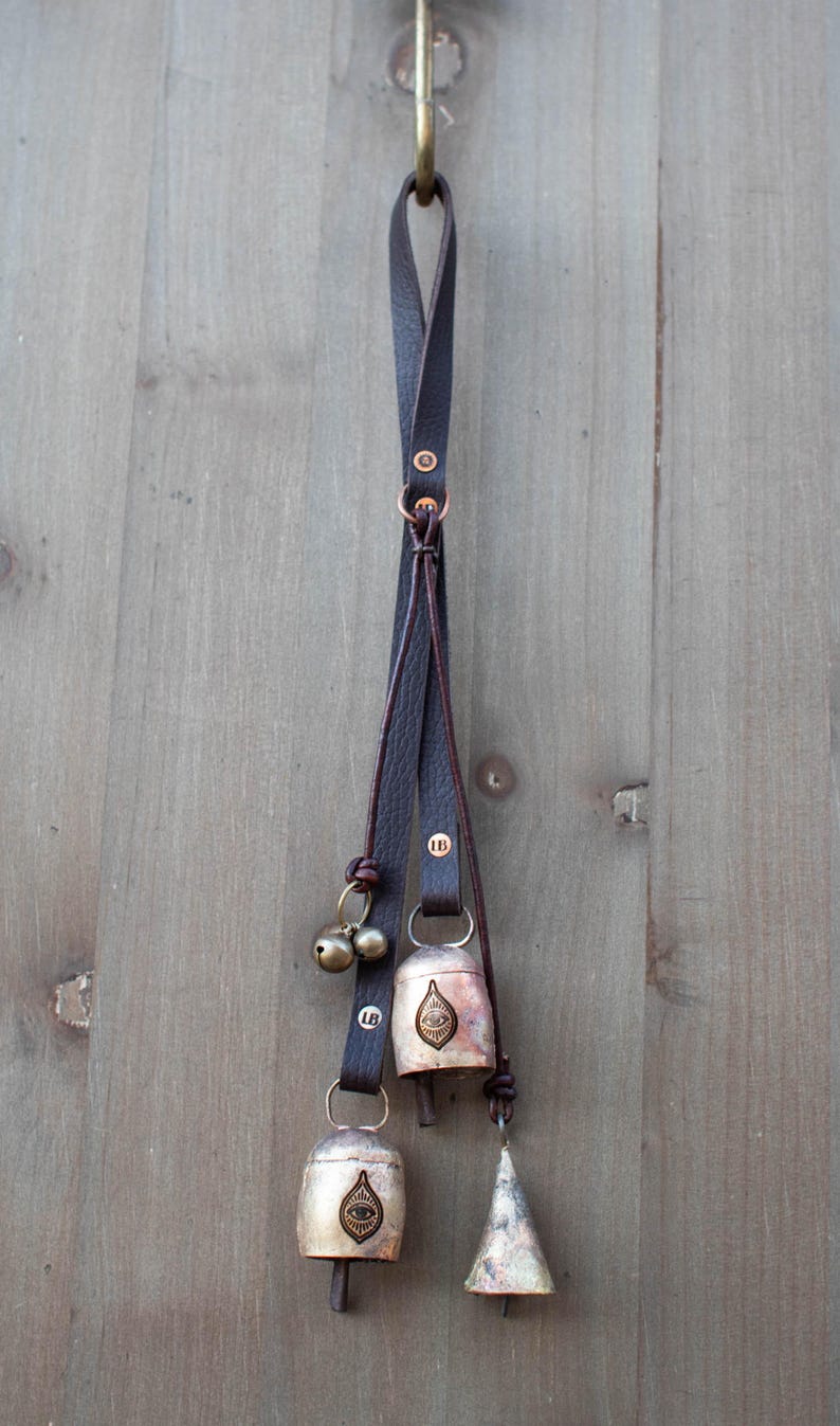 May include: A decorative hanging with three bells and two smaller brass bells. The bells are attached to a dark brown leather strap. The bells have a metallic finish with an eye design. The hanging is suspended from a brass hook.