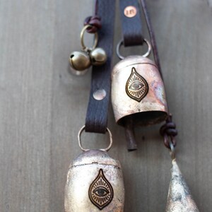 May include: Close-up of three metal bells with leather straps. The bells are in varying shapes, including a cone and a rounded bell shape. Each bell has a unique design, including an eye symbol. The leather straps are brown.