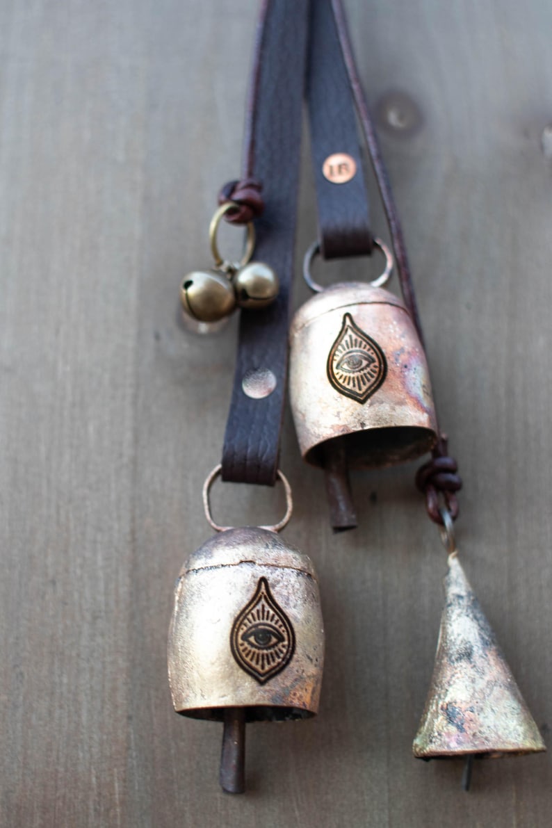May include: Close-up of three metal bells with leather straps. The bells are in varying shapes, including a cone and a rounded bell shape. Each bell has a unique design, including an eye symbol. The leather straps are brown.