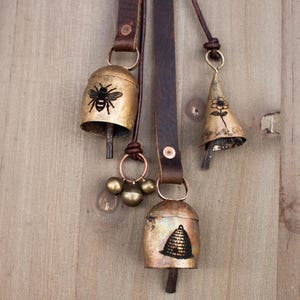 May include: Three rustic metal bells with leather straps hang against a wooden background. The bells are gold-toned with black illustrations of a bee, a flower, and a beehive. The bells are connected with brown leather straps and copper-colored rings.