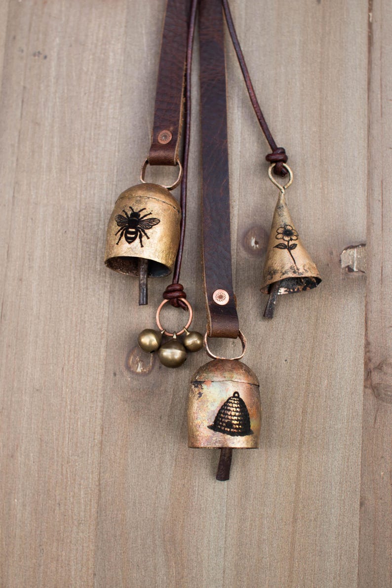 May include: Three rustic metal bells with leather straps hang against a wooden background. The bells are gold-toned with black illustrations of a bee, a flower, and a beehive. The bells are connected with brown leather straps and copper-colored rings.