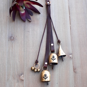 May include: A decorative hanging with three brass bells and a cluster of small gold bells, attached to brown leather straps. The bells have a thistle design. A dark purple flower is in the upper left corner. The background is a light wood surface.