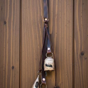 May include: A rustic hanging bell set with a leather strap. The set includes three bells of different shapes and sizes, with a small bell featuring a butterfly design. The bells are suspended against a wooden door.