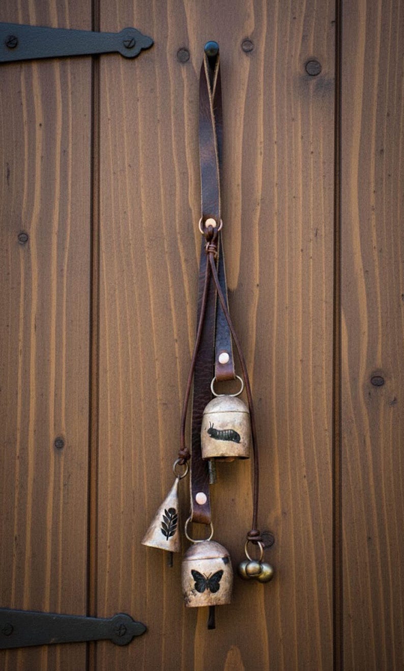 May include: A rustic hanging bell set with a leather strap. The set includes three bells of different shapes and sizes, with a small bell featuring a butterfly design. The bells are suspended against a wooden door.