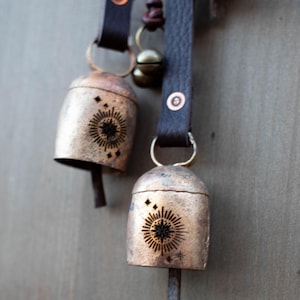May include: Two rustic metal bells with a sunburst design and leather straps. The bells are a tarnished gold color, with a dark sunburst pattern etched into the front. The leather straps are dark brown.