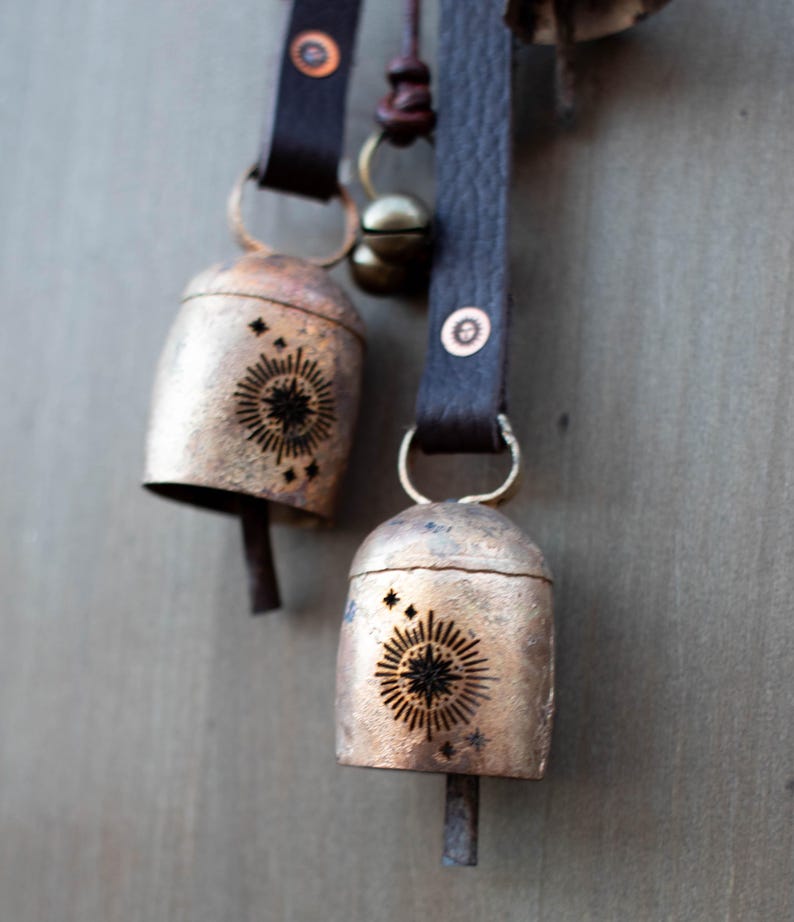 May include: Two rustic metal bells with a sunburst design and leather straps. The bells are a tarnished gold color, with a dark sunburst pattern etched into the front. The leather straps are dark brown.
