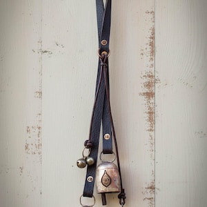 May include: Decorative hanging bells with a dark leather strap. The bells are silver-toned with an embossed design. A small, silver-toned conical bell hangs below. The bells are suspended from a brass hook.