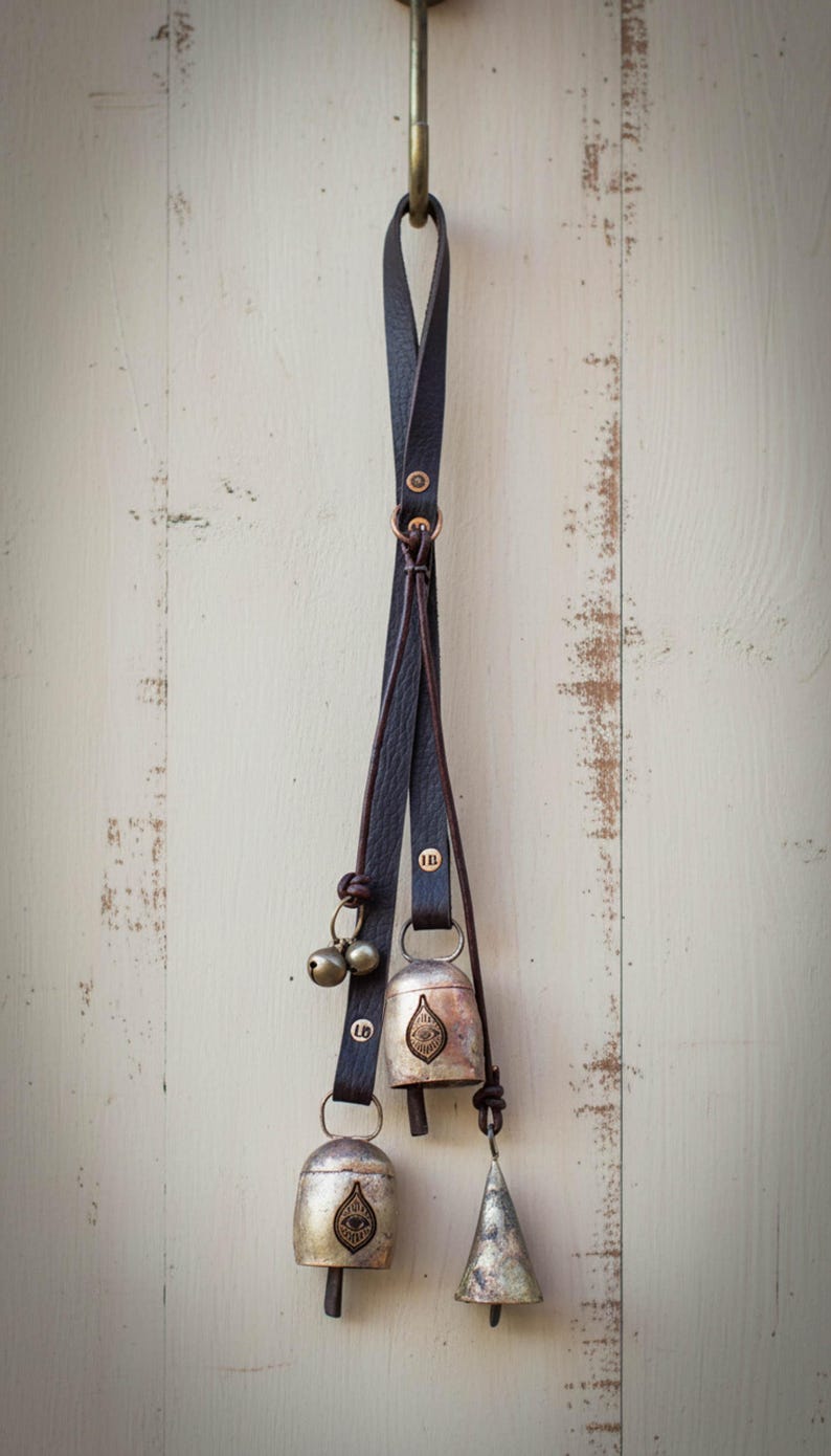 May include: Decorative hanging bells with a dark leather strap. The bells are silver-toned with an embossed design. A small, silver-toned conical bell hangs below. The bells are suspended from a brass hook.