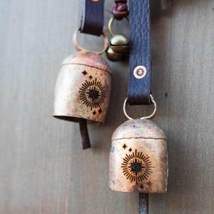 May include: Close-up of rustic metal bells with a sunburst design and star accents. The bells hang from dark brown leather straps with metal rivets. The bells are a warm copper color, and the design is etched in black.