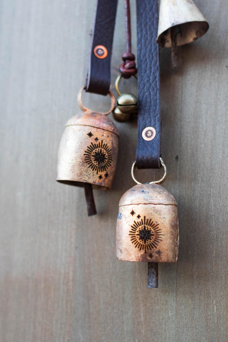 May include: Close-up of rustic metal bells with a sunburst design and star accents. The bells hang from dark brown leather straps with metal rivets. The bells are a warm copper color, and the design is etched in black.