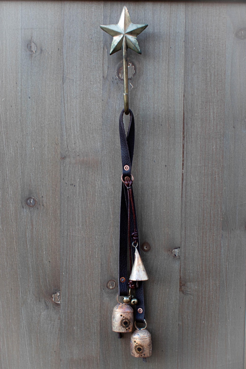 May include: A rustic star-shaped metal hook with a dark leather strap and hanging bells. The bells are various sizes and shapes, with a distressed finish. The hook is mounted on a weathered wooden surface.