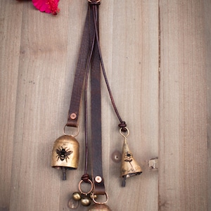 May include: Rustic metal bells with bee and flower designs hang from a brown leather strap. The bells are attached with copper-colored hardware. A bright pink hibiscus flower rests on a wooden surface.