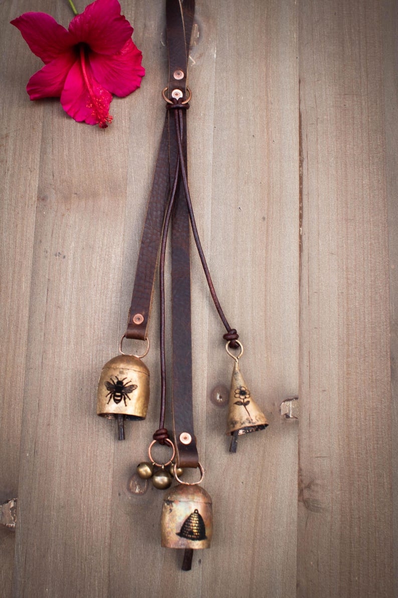 May include: Rustic metal bells with bee and flower designs hang from a brown leather strap. The bells are attached with copper-colored hardware. A bright pink hibiscus flower rests on a wooden surface.